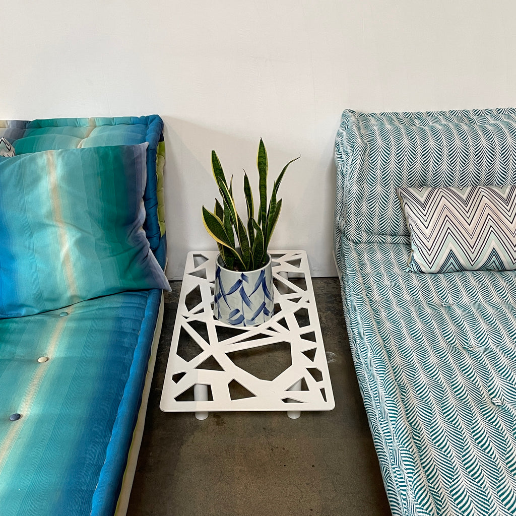 A colorful cushioned bench with stacked pillows next to a Roche Bobois Mah Jong Outdoor Side Table on a wooden floor.