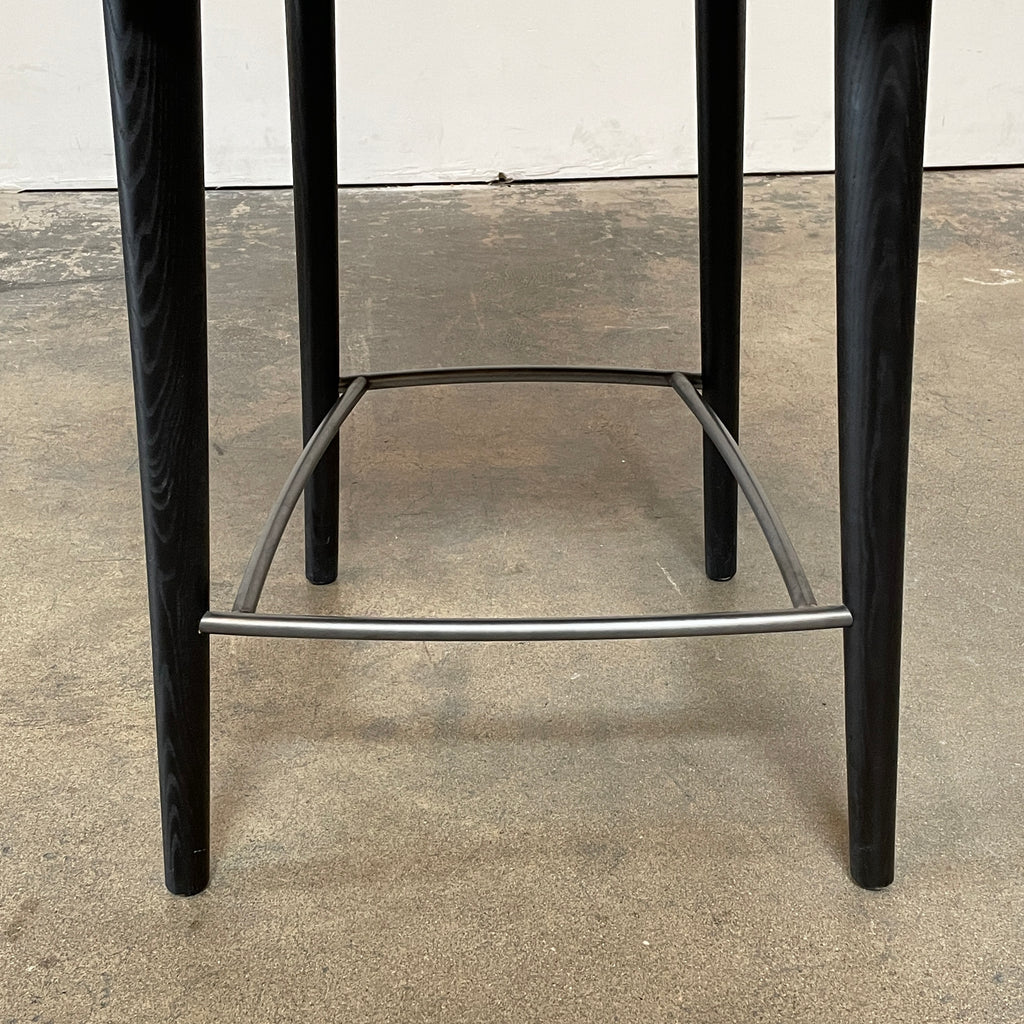 A pair of Minotti Aston Counter Stools, featuring beige cushioned backs and seats supported by sleek black-finished solid ash legs, is displayed against a plain background.