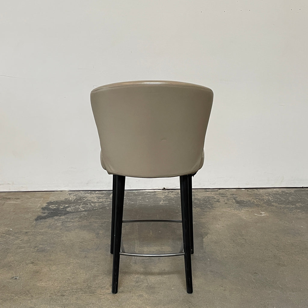 A pair of Minotti Aston Counter Stools, featuring beige cushioned backs and seats supported by sleek black-finished solid ash legs, is displayed against a plain background.