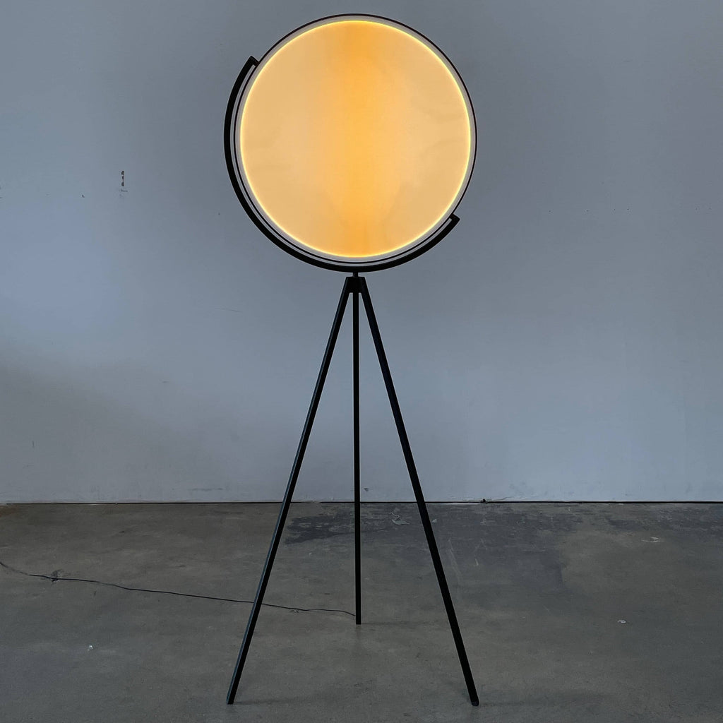 A large circular mirror set in a black metal frame on a tripod stand, isolated against a white background, exudes sophistication akin to the Flos Superloon Floor Light by Flos, providing broad and diffused light that can be dimmed for any ambiance.