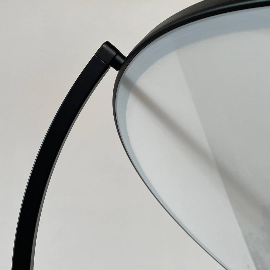 A large circular mirror set in a black metal frame on a tripod stand, isolated against a white background, exudes sophistication akin to the Flos Superloon Floor Light by Flos, providing broad and diffused light that can be dimmed for any ambiance.