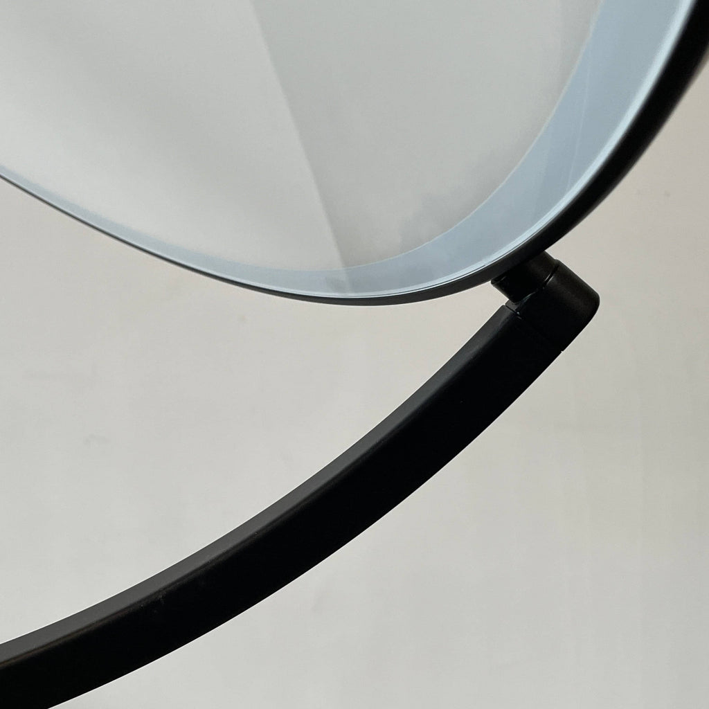 A large circular mirror set in a black metal frame on a tripod stand, isolated against a white background, exudes sophistication akin to the Flos Superloon Floor Light by Flos, providing broad and diffused light that can be dimmed for any ambiance.