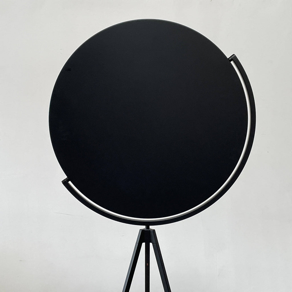 A large circular mirror set in a black metal frame on a tripod stand, isolated against a white background, exudes sophistication akin to the Flos Superloon Floor Light by Flos, providing broad and diffused light that can be dimmed for any ambiance.