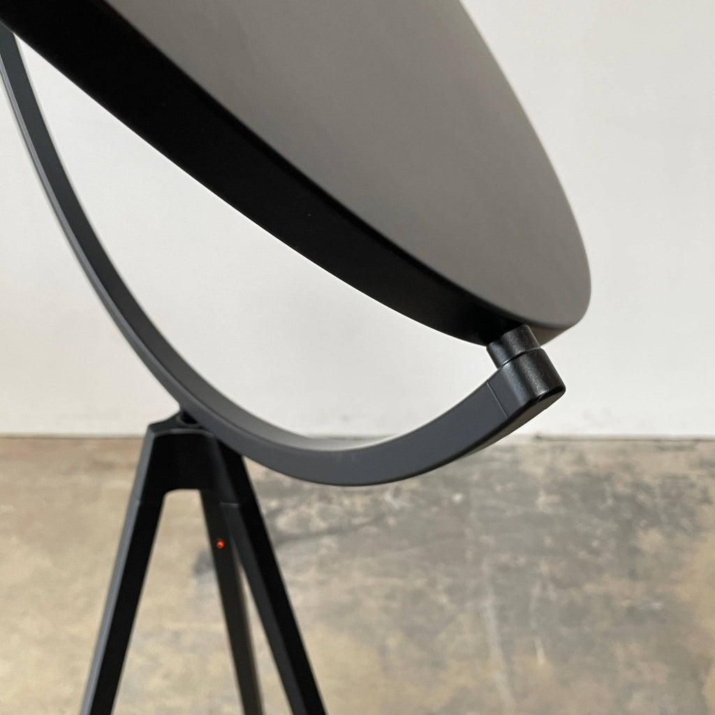 A large circular mirror set in a black metal frame on a tripod stand, isolated against a white background, exudes sophistication akin to the Flos Superloon Floor Light by Flos, providing broad and diffused light that can be dimmed for any ambiance.