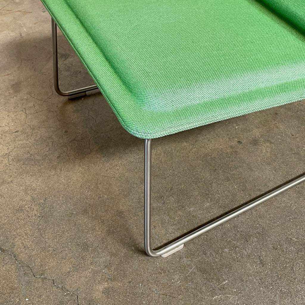 A Cappellini Low Pad Lounge Chair with a metal frame, reminiscent of Jasper Morrison's design, sits next to a small, round side table holding a cup and saucer, a notebook, and a pen. The setting is a minimalist indoor space with a concrete floor.