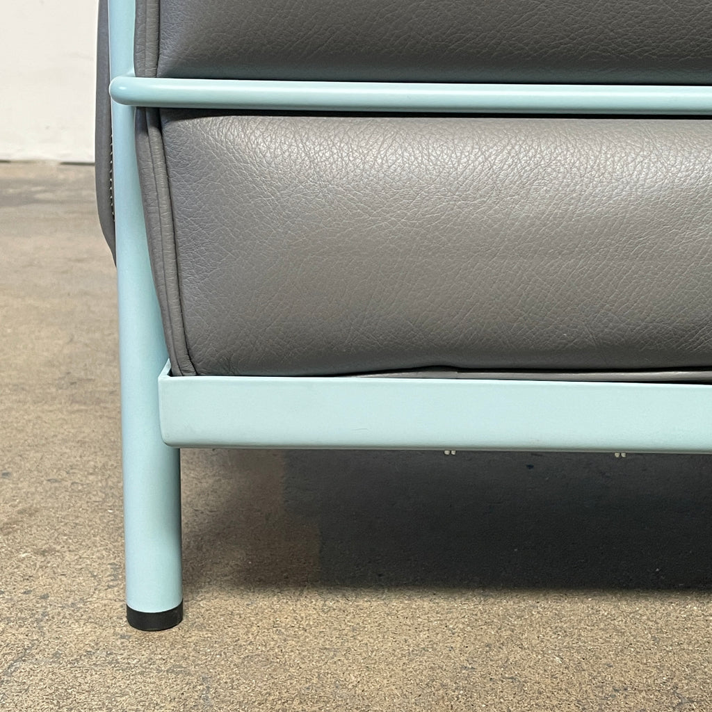 Two gray leather Cassina LC2 Lounge Chairs with light blue metal frames, designed by Cassina. One chair faces forward while the other reveals its back view, adorned with a red tag for a pop of color.