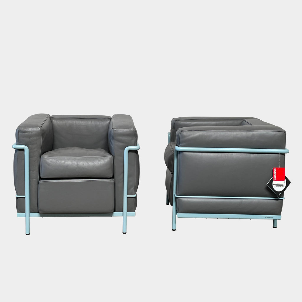 Two gray leather Cassina LC2 Lounge Chairs with light blue metal frames, designed by Cassina. One chair faces forward while the other reveals its back view, adorned with a red tag for a pop of color.