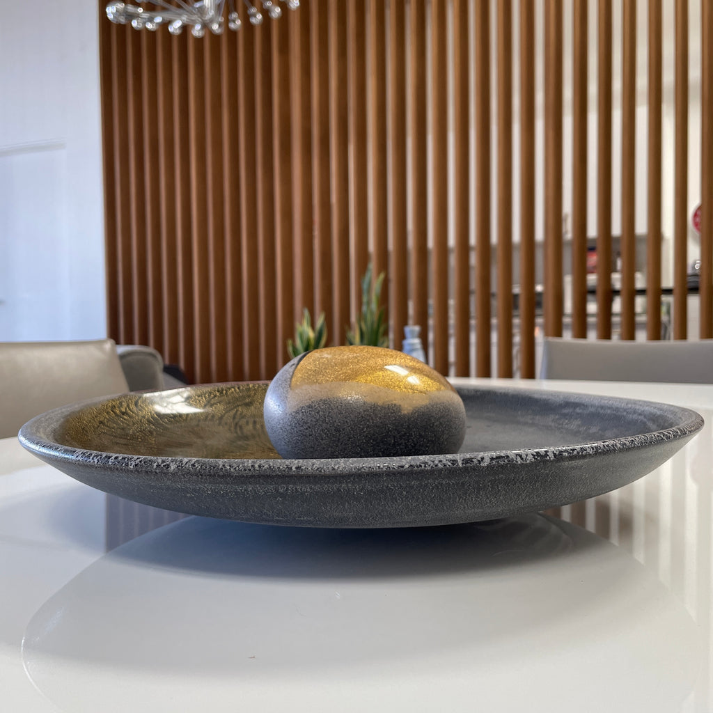 A round ceramic plate featuring a central raised circular design, predominantly gray with earthy bronze tones and textured streaks on one side, reminiscent of the artistry seen in the Alfredo Barbini Murano Glass Plate And Object.