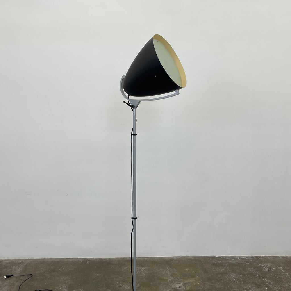 The Pallucco Italia Faro Floor Lamp, featuring a round, cone-shaped shade and set on a square base, exudes industrial design in a minimal room with a concrete floor.