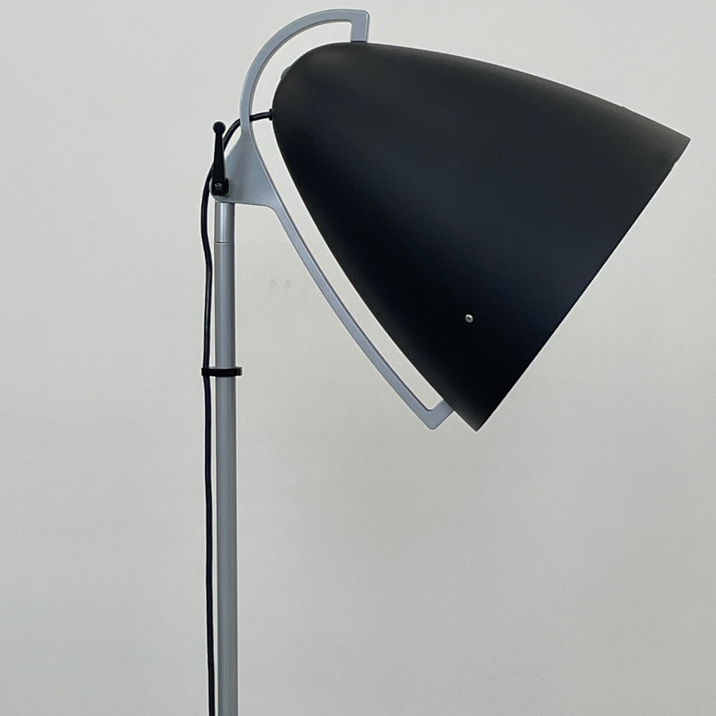 The Pallucco Italia Faro Floor Lamp, featuring a round, cone-shaped shade and set on a square base, exudes industrial design in a minimal room with a concrete floor.