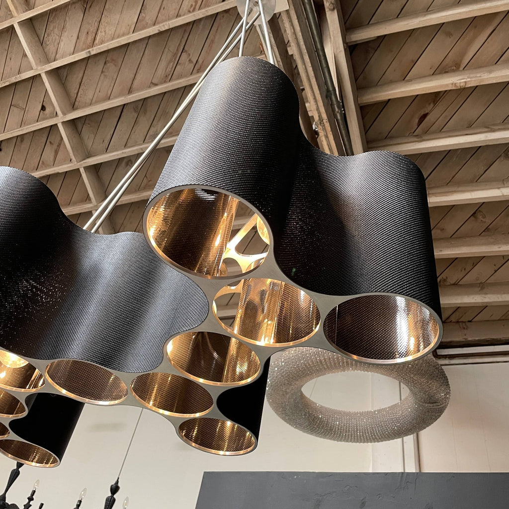 A modern metal chandelier with multiple cylindrical shades connected by thin, curved attachments, hanging from a ceiling mount—a design reminiscent of the Ralph Pucci Linear Cumulus Ceiling Light.